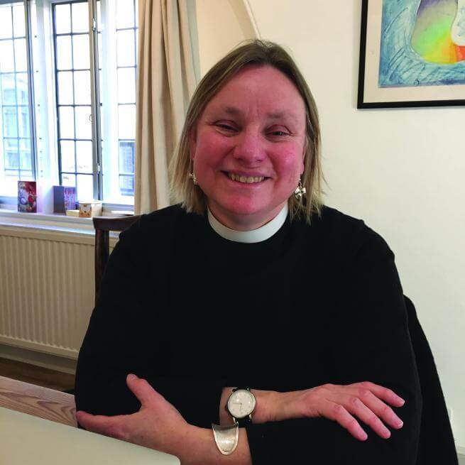 Jessica Martin is a white woman with short blond hair, photographed in clerical shirt and in her house, sitting leaning on a table and smiling.