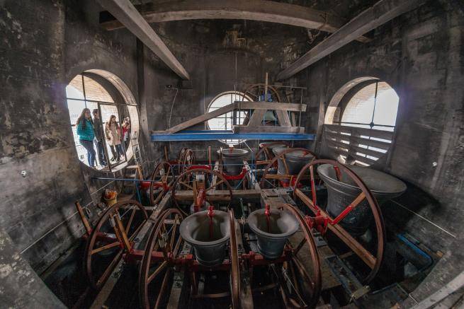 A wide angled shot from inside the north west tower, showing the 12 change ringing bells as they are ringing. Some children stand to one side outside the tower, on the roof, looking in.