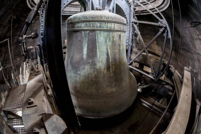 A huge cast bell, stained green in places, held in place by a large iron structure, inside a stone building