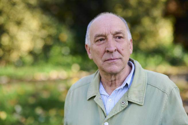 Bob Gilbert is a white man with bald head, wearing a pale green jacket over a blue shirt. He stands outdoors with sun dappled greenery in the blurred background.