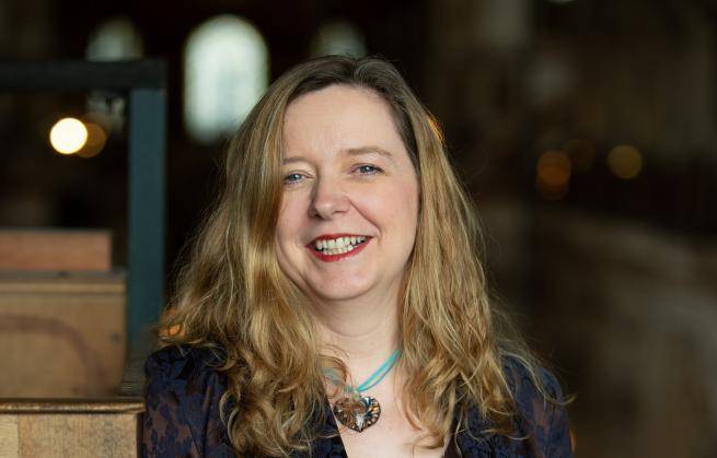 Paula Gooder is a white woman with long blond hair wearing a dark jacket and blue pendant necklace. She smiles to the camera and is standing indoors with the blurry glow of windows behind her in what appears to be a very large space.