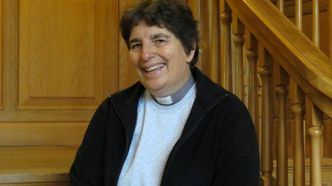 Paula Hollingsworth, a white woman with short dark hair in a clerical collar, stands in front of a wooden staircase.