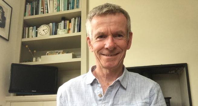 Christopher Chapman is a white man with short grey hair wearing a pair grey casual shirt and sitting in front of a bookcase, cupboard and fireplace.