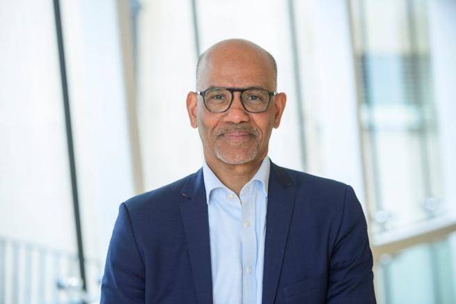 Alan Smith, a black man with bald head and black glasses wearing a navy blazer over a pale blue shirt, stands in front of large glass windows.