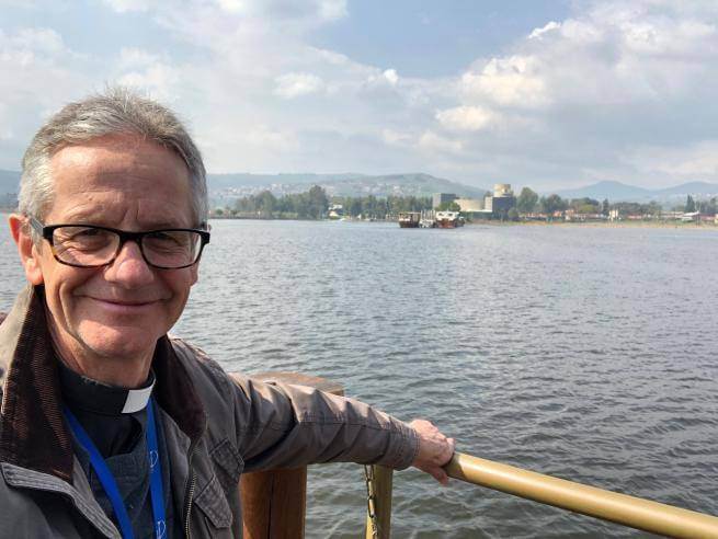 Richard is a white man with short grey hair and black rimmed glasses. He wears a clerical collar and black shirt under a casual light jacket and stands holding onto the wooden railing of a boat or a jetty on a lake. The shoreline in the distance has trees, and some buildings, and there are mountains in the distant background. The sky is pale blue with some clouds and the sun is shining on Richard.
