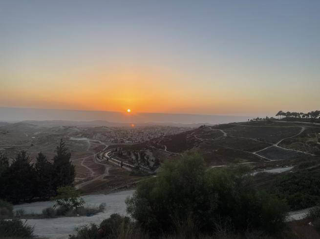 A view over the Jordan Valley from the Mount of Olives, looking down towards a distant town with the sun rising in an orange-tinged sky in the distance.