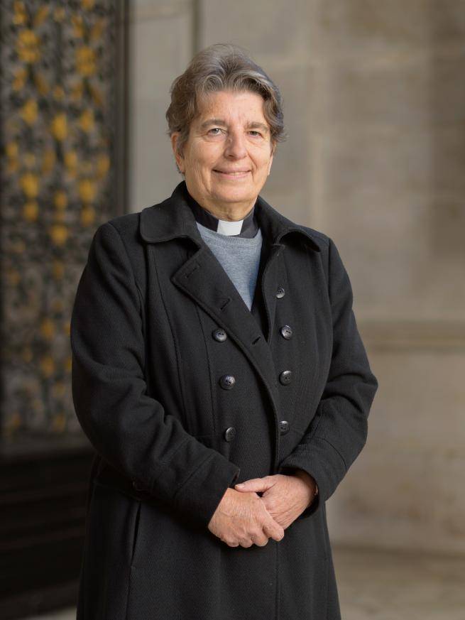 Paula is a white woman with short dark grey hair and wears a clerical collar under a grey shirt and black double breasted coat. 