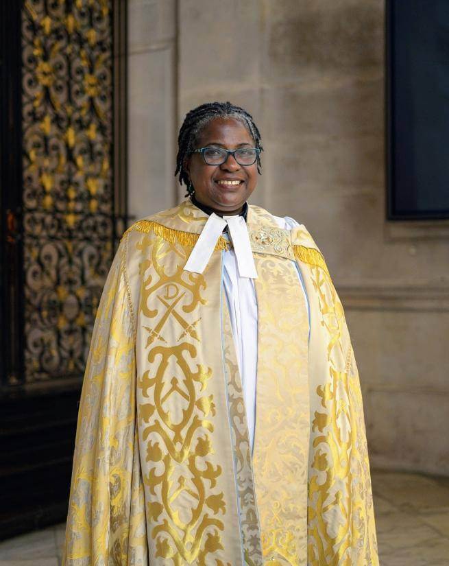 Revd Canon Adéolá is a black woman with short dark hair and glasses. She wears a golden yellow embroidered cope and stands before a portland stone wall and an intricately designed gold and black metal gate.