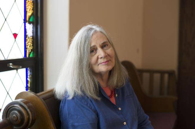 Marilynne is a white woman with long, straight grey hair. She wears a coral pink t-shirt and necklace under a dark blue shirt and sits on a dark wooden pew before a window with stained glass panels. 