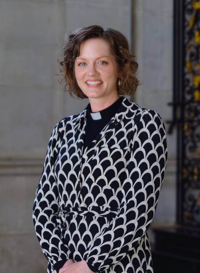 Tessa is a young white woman with wavy brown hair wearing a clerical collar under a black and white patterned dress, standing by the wrought iron gates near the high altar at St Paul's.