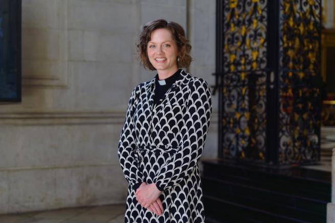 Tessa is a young white woman with wavy brown hair wearing a clerical collar under a black and white patterned dress, standing by the wrought iron gates near the high altar at St Paul's.