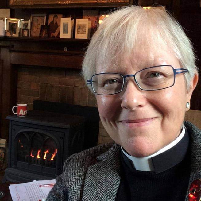 Cally is a white woman with white hair and glasses wearing a clerical collar under a black shirt and grey tweed jacket. She is in front of a mantlepiece decorated with card, framed photos and an Icon, and a lit log burner. She is smiling.