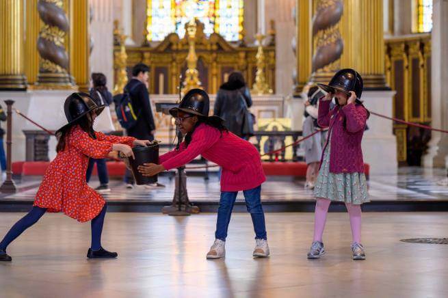 Three young children pass a bucket to one another, wearing fire helmets, role playing firefighting during the Great Fire of London.