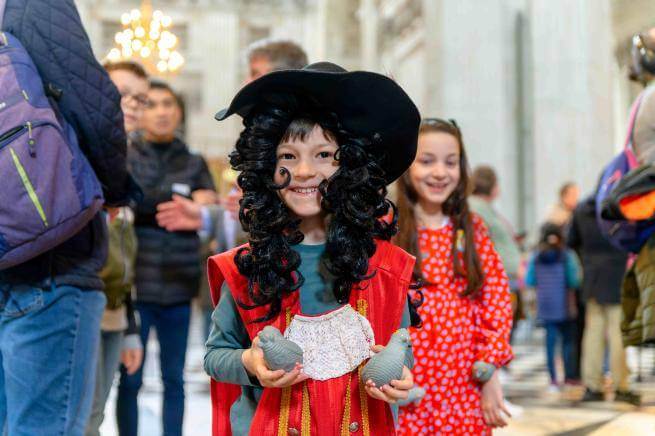 A young child wearing a long black, curly wig and wide brimmed hat, dressed as Christopher Wren.