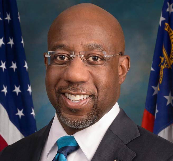 Raphael Warnock is a black man with a bald head, wearing frameless glasses, a dark suit with a white shirt and stripy tie. Behind him are two US flags.