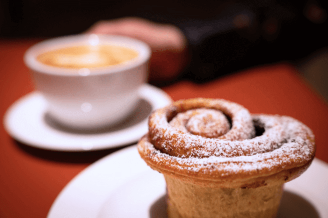 A close up shot of a cinnamon bun dusted with icing sugar in the foreground, and a cup of coffee in the background.