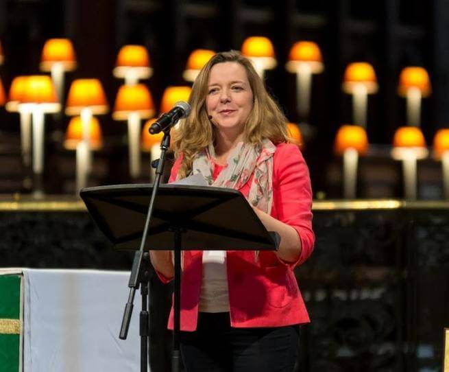 Paula is a white woman with long blond hair wearing a red jacket and grey scarf and standing in front of a microphone and music stand with the glowing lamps of the quire of St Paul's Cathedral in the background