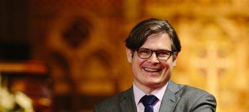 Andreas is a white man with short dark hair and glasses wearing a grey suite, pink shirt and dark tie, with the golden stonework of a Cathedral interior behind him.