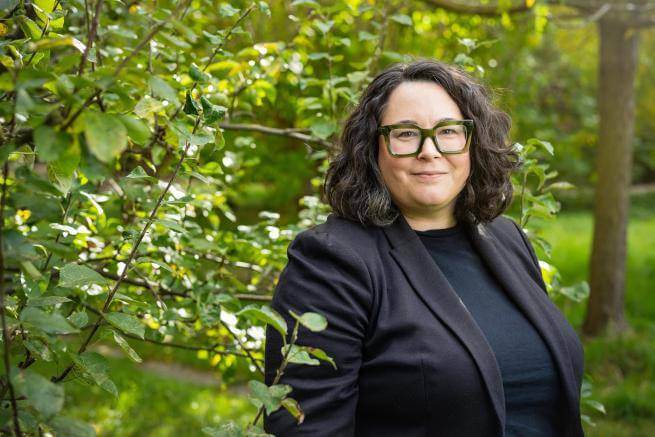 A white woman in clerical collar wearing dark green glasses standing in front of sun dappled trees.