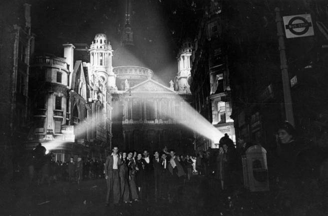 The West End of St Paul's, lit by searchlights, with a crowd of people posing for a photo in front of it.