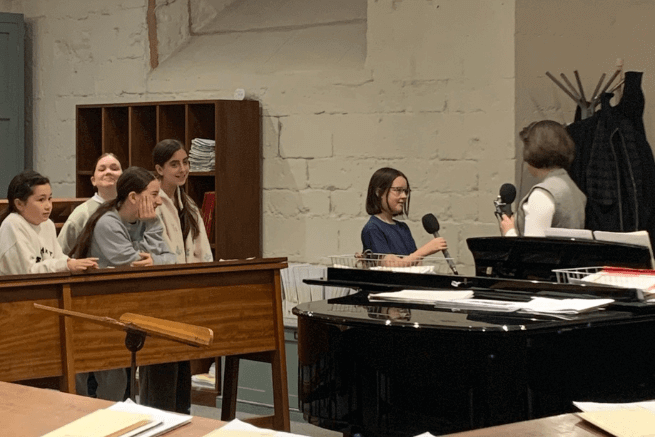 A girl chorister is interviewed by a BBC presenter in the choir practice room, while other choristers look on.