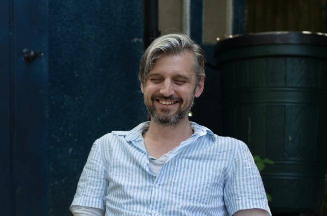 David is a white man with light brown hair and a darker beard, sitting outside in a garden chair, wearing a pale blue short sleeved shirt.