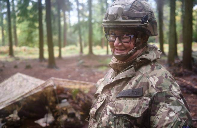 Adele wears army fatigues and a helmet and stands in front of a shelter in woodland. She wears a badge which says Padre Rees.