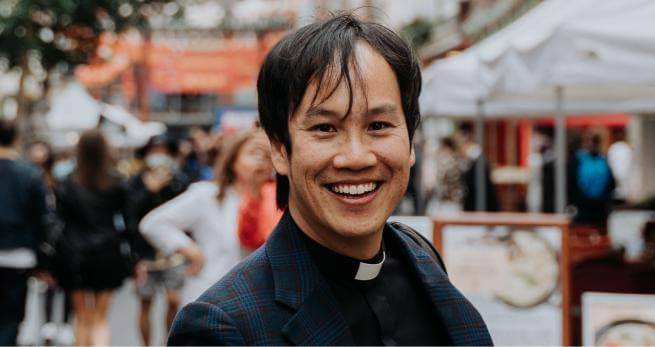 Mark Nam headshot wearing a clerical collar under a black suit and shirt, standing in a busy marketplace