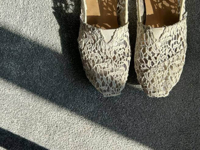 a pair of white embroidered slippers, worn but with lots of life left in them, on concrete in sunshine with a shadow falling across their tips