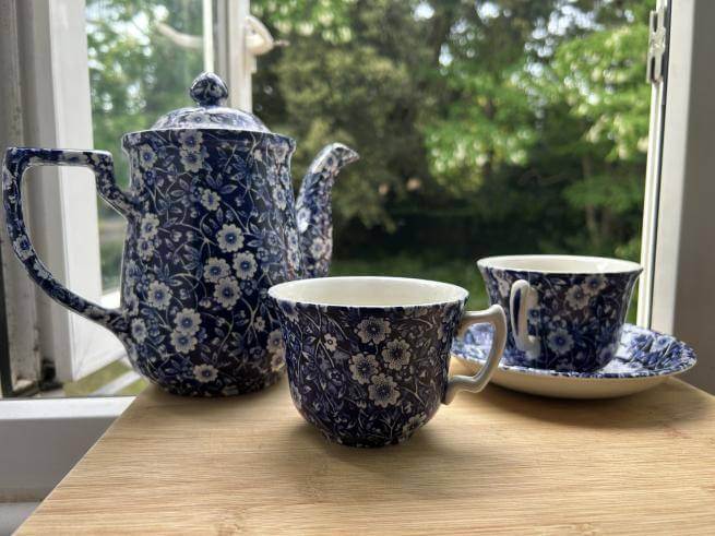A blue and white calico tea set sits on a wooden table by and open window overlooking a garden