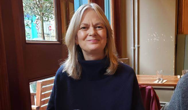 Linda wears a navy polo neck jumper and sits in a cafe by windows overlooking a tree in a city