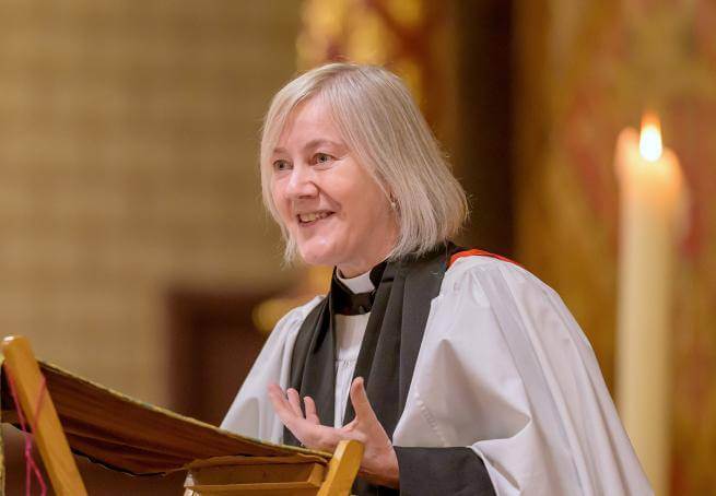 Ellen wears a clerical collar and robes while standing at a lectern, her hand outstretched as she talks