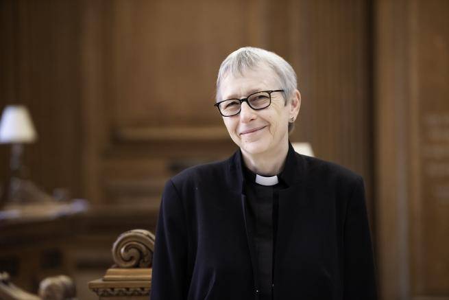 Alison wears a black jacket and shirt with a clerical collar and is photographed inside a church
