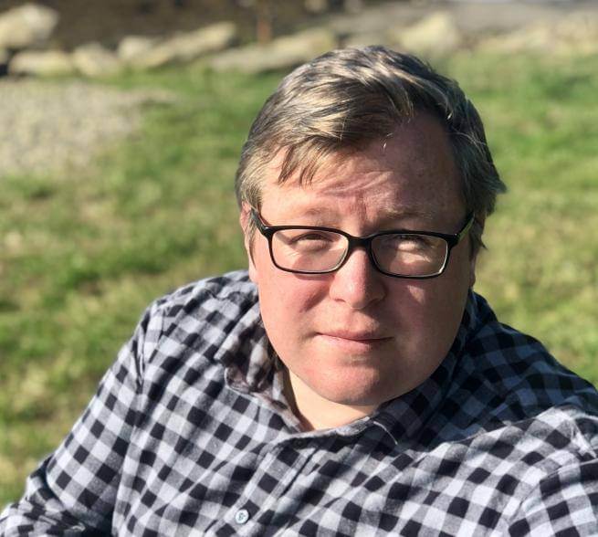 A man with blond hair and glasses wearing a checked shirt