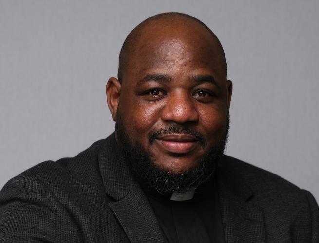 Carlton wears a clerical collar with a black shirt and jacket