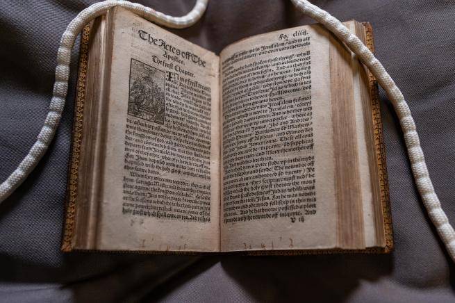 Tyndale Bible open at "The Acts of the Apostles". Long white "snake" weights are visible holding the top right and left corners of paper open. 