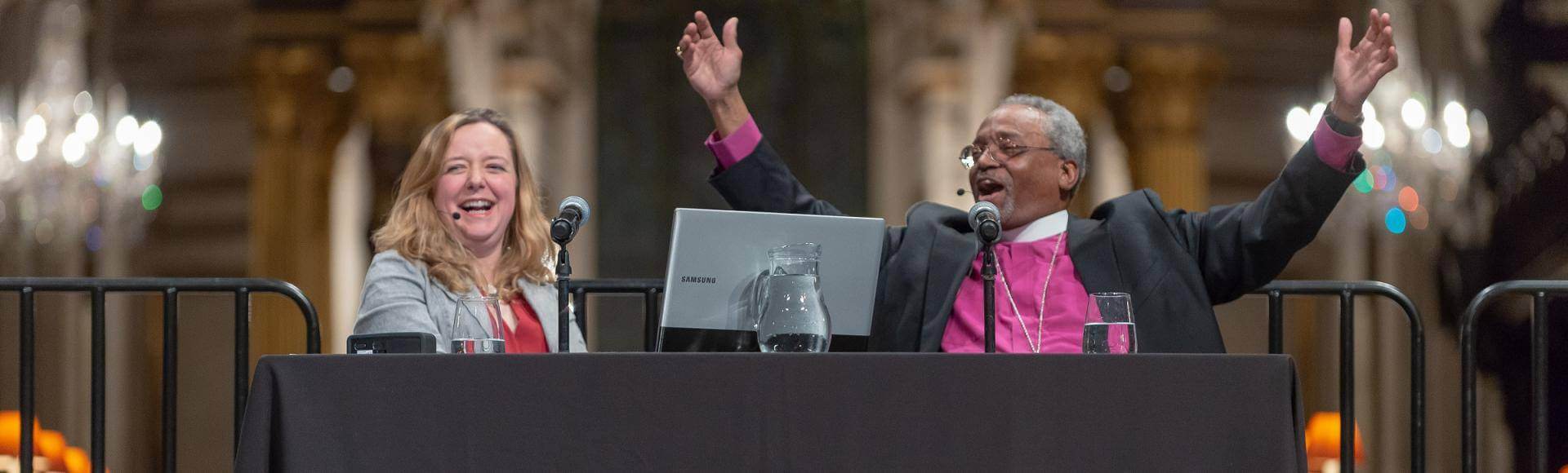 Bishop Curry raises his arms and Paula Gooder laughs alongside on the stage at an event at St Paul's Cathedral