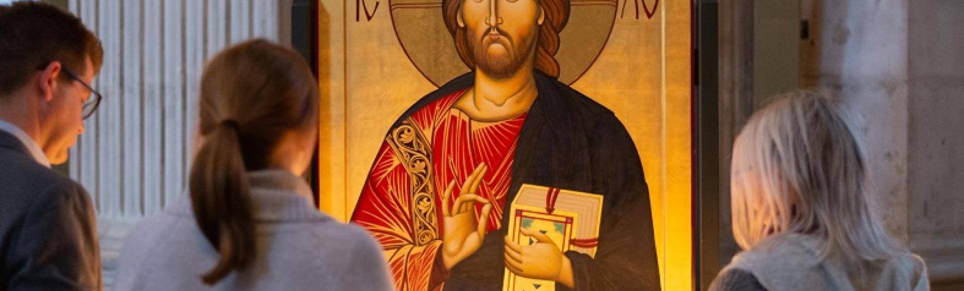 Two women and a man stand before an Icon of Jesus and light candles as they reflect.