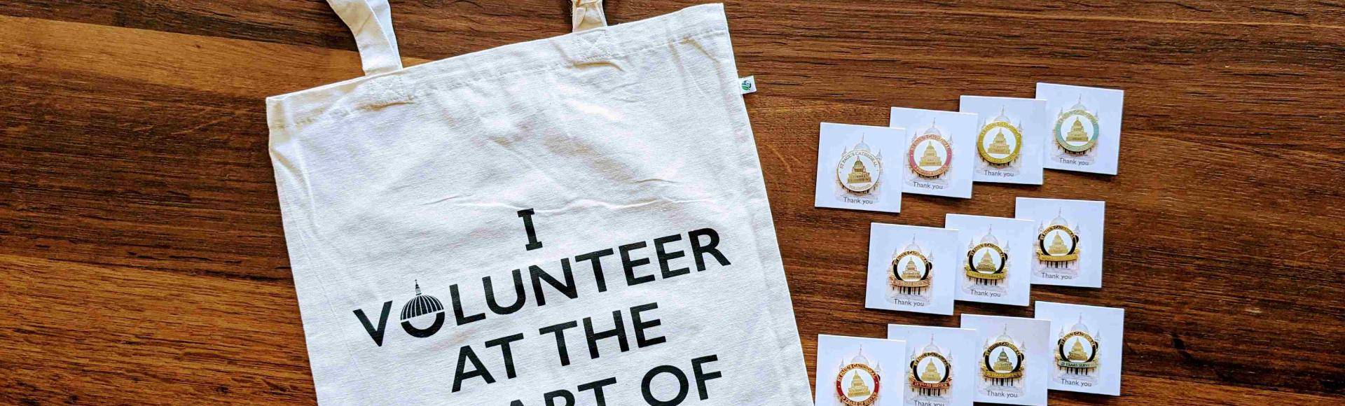 A white cotton tote bag laid flat on a wooden table. The words 'I volunteer at the heart of the City' are printed in black lettering on the bag. Beside the bag are 11 metal pin badges featuring the dome of St Paul's.