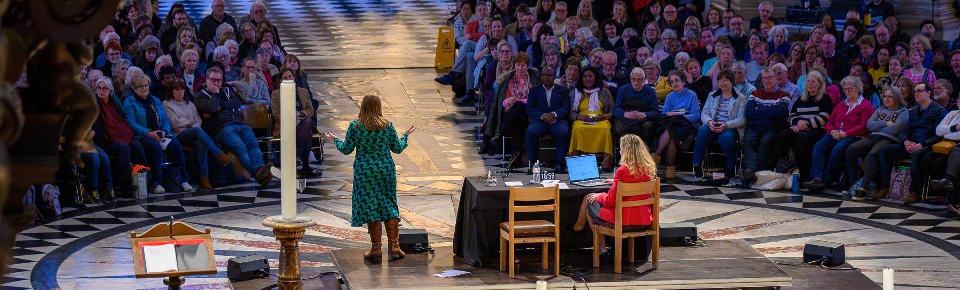 A view of the cathedral with a large audience facing a stage with a speaker in a green dress