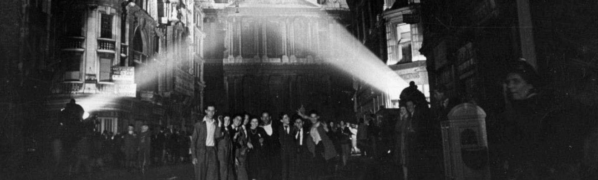 The West End of St Paul's, lit by searchlights, with a crowd of people posing for a photo in front of it.