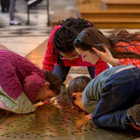 Four young children kneel on the floor of St Paul's Cathedral, with their faces close to the ground, looking through a golden grille through to the Cathedral Crypt.