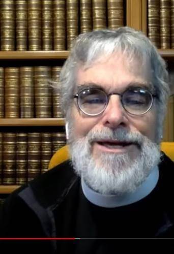 Brother Guy is a white man with round glasses, grey hair and a full grey beard sitting in front of bookshelves full of bound leather books., and a globe behind to his left.
