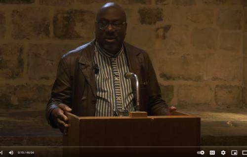 A photo taken from the film of Anthony Reddie speaking in the Wren Suite at St Paul's Cathedral. Pictured standing at a lectern in front of a wall of exposed brickwork, he is a Black man wearing a striped shirt and a brown leather jacket.