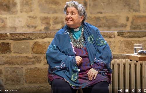 June Boyce-Tillman is a white woman with grey hair gathered in a bun, wearing a turquoise blue clerical shirt under a blue shawl. She is pictured sitting indoors in front of an exposed brick wall.