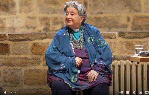 June Boyce-Tillman is a white woman with grey hair gathered in a bun, wearing a turquoise blue clerical shirt under a blue shawl. She is pictured sitting indoors in front of an exposed brick wall.