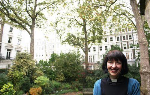 Carys, a white woman with short brown hair wearing a clerical collar, stands in a garden square in a city