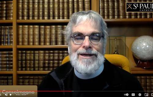 Brother Guy is a white man with round glasses, grey hair and a full grey beard sitting in front of bookshelves full of bound leather books., and a globe behind to his left.