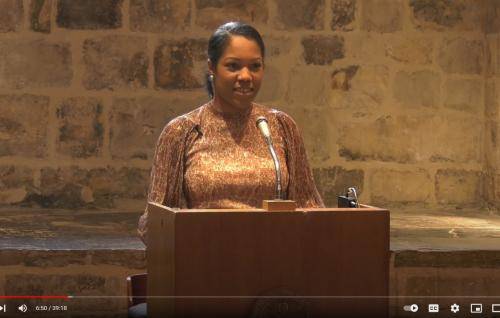 Selina Stone is a young black woman and is pictured at the lectern in the Wren Suite, an exposed brick wall behind her.
