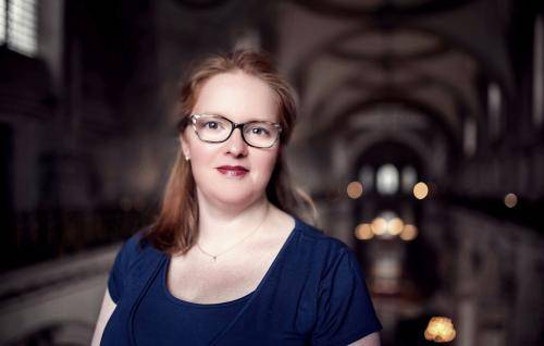 Carris, a white woman with auburn hair and glasses, stands in the cathedral, the lights of the nave and the decorative ceiling blurred in the background behind her.